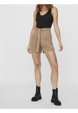 Kvinde iført en sort ærmeløs top, beige højtaljede shorts med bælte og sorte ankelstøvler, stående mod en ensfarvet baggrund.