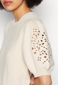 Beige svetr s balónovými rukávy a květinovými výřezy. Žebrovaný výstřih a manžety s jemnou texturou.