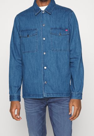 Mand iført blå denimjakke med knaplukning foran og to brystlommer med klapper, kombineret med blå jeans.