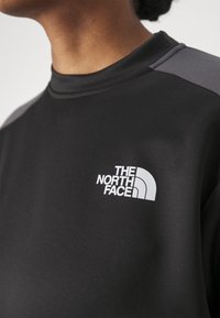 Mustat urheilu-topit, jossa on korkea kaulus, harmaat yksityiskohdat harteilla ja valkoinen "The North Face" -logo. Sujuva pinta.