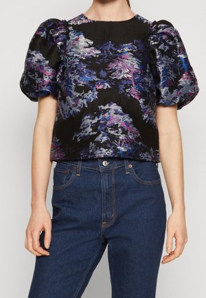 Musta pusero, jossa on pöyhistetyt lyhyet hihat ja violettiin ja siniseen vivahtava abstrakti kukkakuosi, yhdistetty tummansinisiin korkea vyötäröisiin farkkuihin.