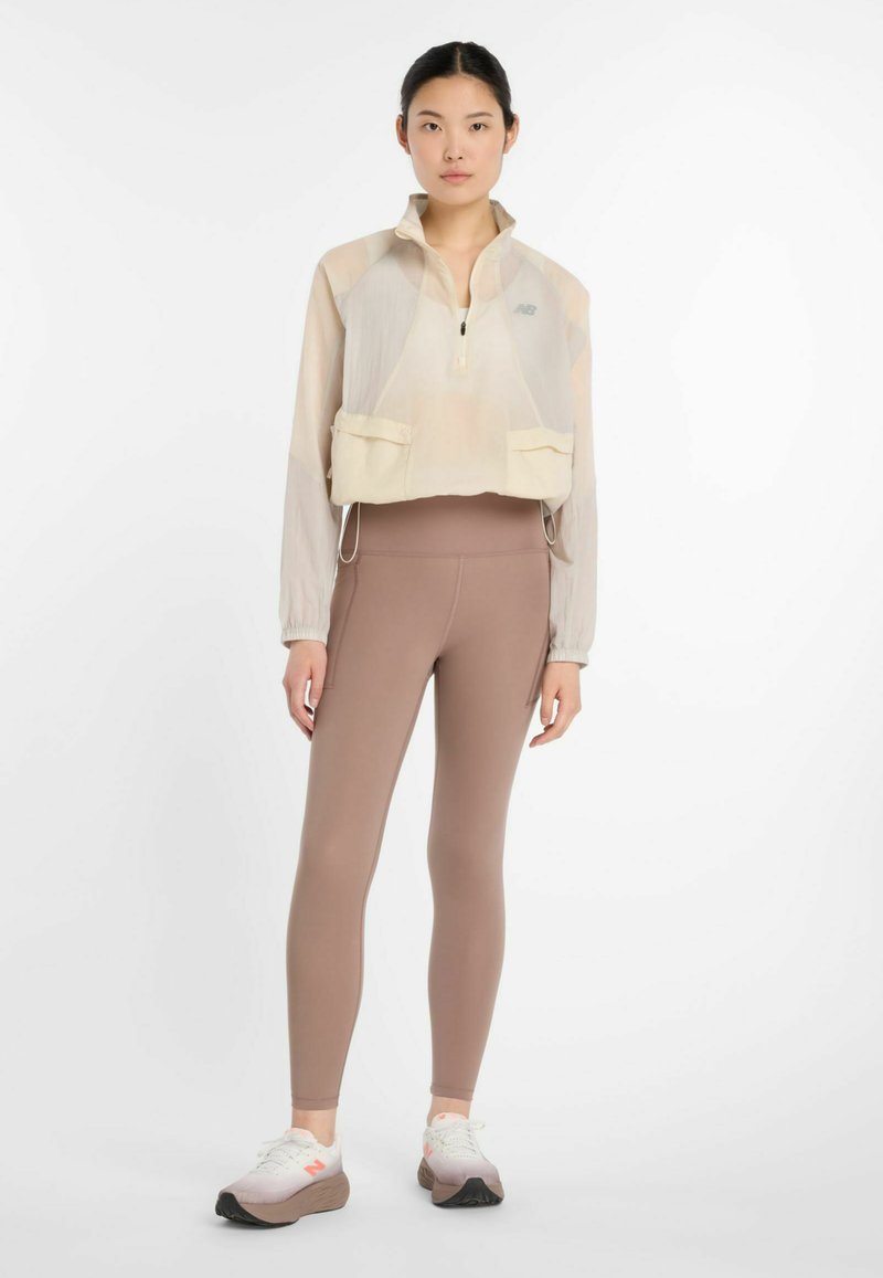 Gekürzte hellbeige Jacke mit zwei Vorderaschen, Reißverschluss und elastischen Bündchen, kombiniert mit eng anliegenden braunen Leggings und weißen Turnschuhen.