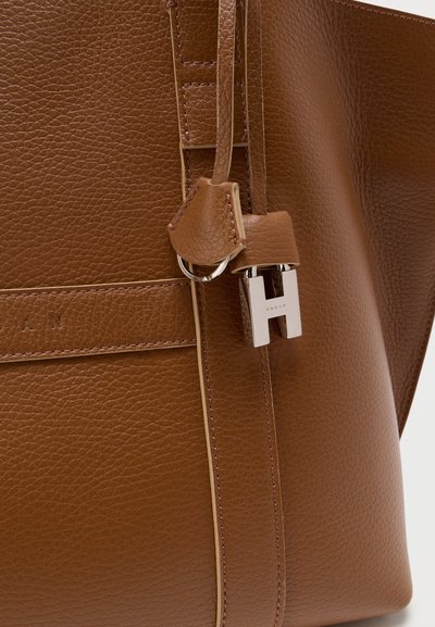 Gros plan sur un sac en cuir marron texturé avec une fermeture "H" argentée et un détail de sangle en cuir.