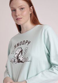 Camisa de manga larga verde menta hecha de tela suave, con un gráfico de Snoopy que presenta personajes de dibujos animados durmiendo, contorneados en marrón. Cuello redondo.