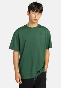 T-shirt verde in cotone con maniche corte, scollatura a girocollo e un piccolo logo stampato sul petto, abbinato a jeans scuri.