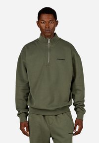 Olīvu zaļš sporta krekls ar ceturtdaļas rāvējslēdzēju, ribotām piedurknēm un apakšmalu. Uz krūtīm ir melns "PEGADOR" logo. Gluda tekstūra.