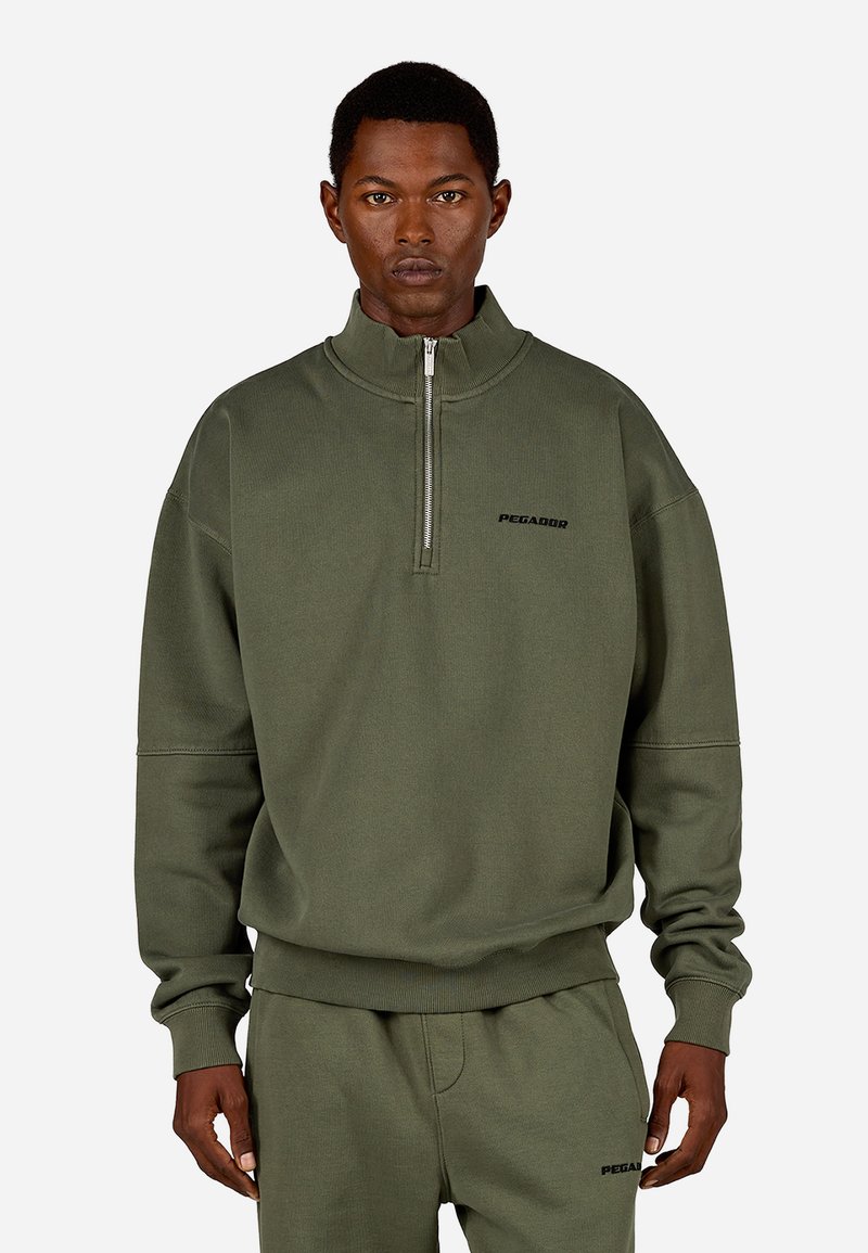 Olīvu zaļš sporta krekls ar ceturtdaļas rāvējslēdzēju, ribotām piedurknēm un apakšmalu. Uz krūtīm ir melns "PEGADOR" logo. Gluda tekstūra.