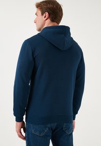 Sudadera azul marino con bolsillo canguro y capucha con cordón, hecha de una tela suave. Presenta puños y dobladillo acanalados, combinada con jeans de mezclilla.