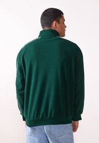 Grøn fleece zip-up jakke med høj krave, hvide sidelængder, ribbede manchetter og kant. Blød tekstur, afslappet pasform og langærmede.