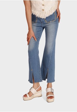 Kvinde iført lyseblå jeans med frontslids og blomsterdekorationer, parret med hvide wedgesandaler og en cremet blondebluse.