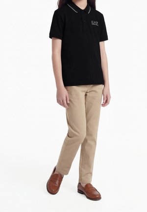 Enfant portant un polo noir EA7, un pantalon beige et des mocassins en cuir marron, debout avec un pied légèrement avancé sur fond blanc.