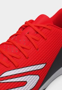 Κοντινό πλάνο ενός κόκκινου αθλητικού παπουτσιού με κόκκινα κορδόνια, διάτρητη γλώσσα, υφή στο επάνω μέρος και λευκό στυλιζαρισμένο λογότυπο στο πλάι.