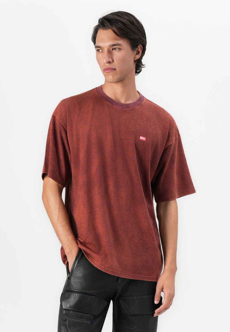 Młody mężczyzna w wyblakłej czerwonej koszulce Diesel i czarnych spodniach cargo, stoi z jedną ręką w kieszeni na tle białej ściany.