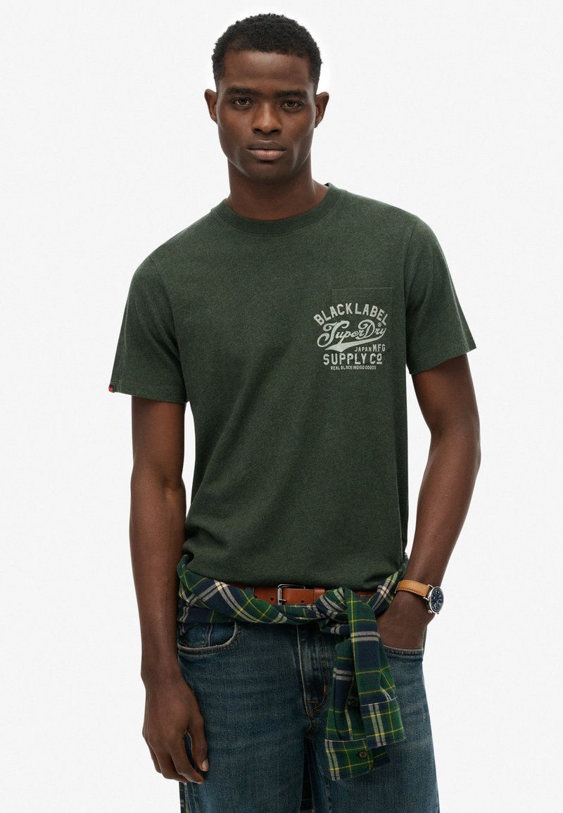 Groene heidelogo t-shirt met korte mouwen en een borstzak met een grafisch logo; gecombineerd met blauwe jeans en een geruit shirt dat om de taille is geknoopt.