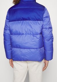 Blå vadderad jacka med en glansig, quiltad yta och horisontella sömmar. Har en hög krage och är vadderad för värme.