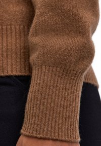 Maglione marrone con texture in primo piano, con polsini a coste e un corpo liscio. Il materiale sembra morbido, con una leggera lucentezza.