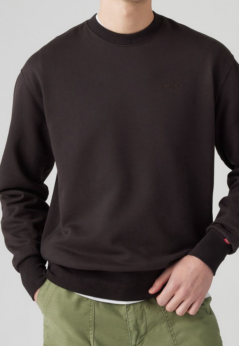 Felpa marrone con collo rotondo, consistenza morbida e discreto marchio Levi's. Presenta polsini e orlo a coste, indossata con pantaloni verdi.