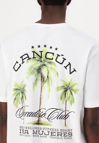 Hvid bomulds t-shirt med palmetegninger, "CANCÚN" tekst i fed skrift og "Paradise Club" i kursiv. Bagside med stjernedesign.