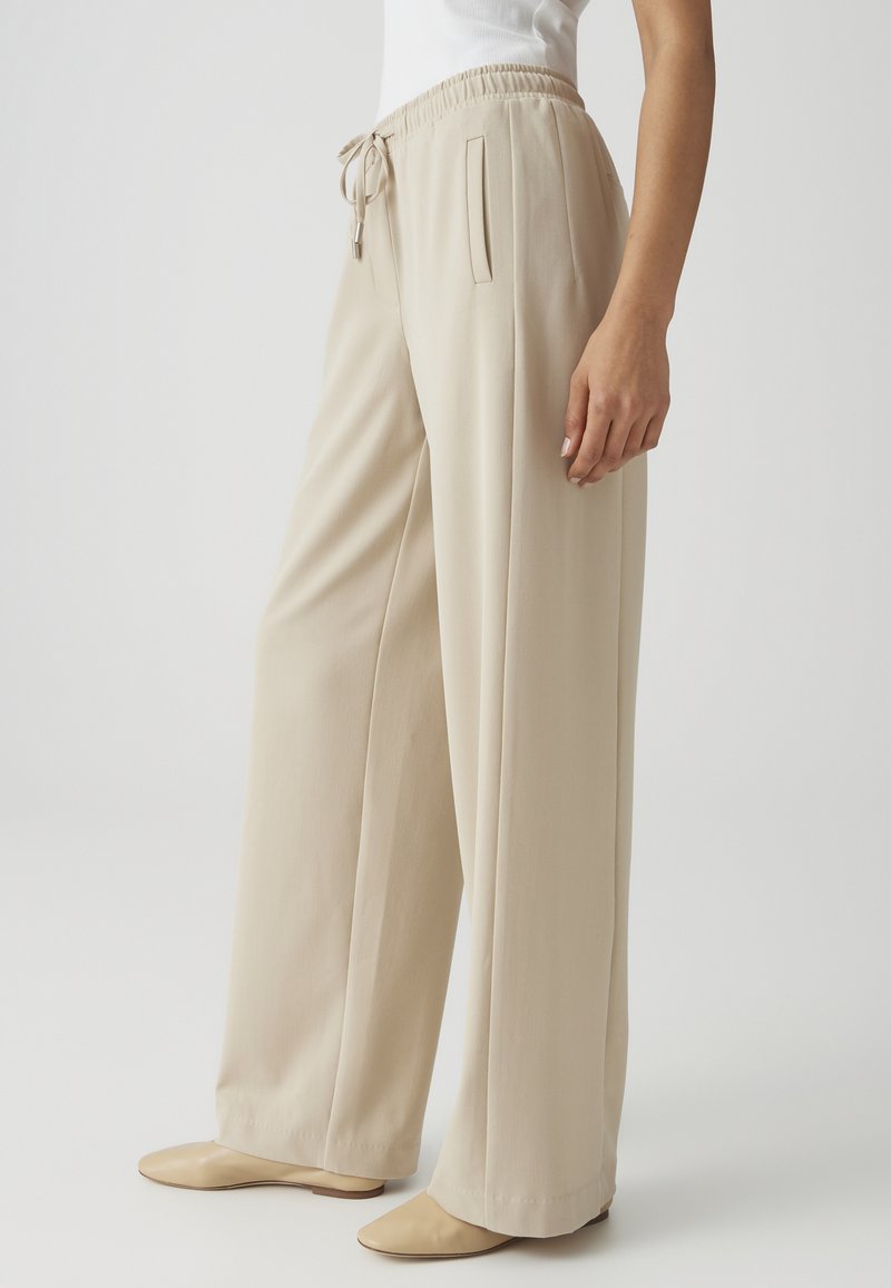 Donna che indossa pantaloni beige a gamba larga con vita elasticizzata e coulisse, abbinati a scarpe beige senza lacci.
