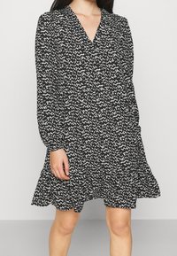 Mekko, jossa on musta-valkoinen kuvio, V-pääntie, pitkät hihat ja röyhelöhelma. Valmistettu kevyestä kankaasta, jossa on valuva muoto.