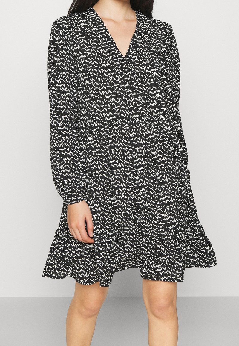 Mekko, jossa on musta-valkoinen kuvio, V-pääntie, pitkät hihat ja röyhelöhelma. Valmistettu kevyestä kankaasta, jossa on valuva muoto.