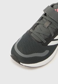 Μαύρο αθλητικό παπούτσι με επάνω μέρος από δίχτυ, γκρι λεπτομέρειες, στρογγυλή μύτη και ελαστικά κορδόνια. Διαθέτει ιμάντα με βέλκρο και λευκή σόλα με σκούρο πέλμα.