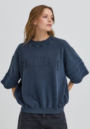 Kvinde med lysebrunt hår iført oversized mørkeblå "Ball Jeans Italy" sweatshirt og mørke bukser, som kigger mod sin højre side.