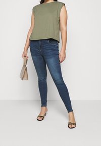 Top corto verde oliva con hombros anchos, combinado con jeans ajustados azul oscuro. Los accesorios incluyen un bolso beige texturizado y sandalias con estampado animal.