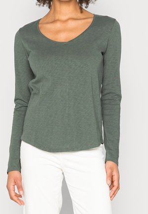 Žena stojící před jednobarevným pozadím, oblečená v khaki zeleném tričku s dlouhým rukávem a výstřihem do V a bílých kalhotách.