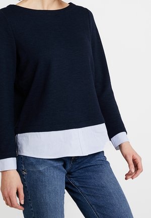 Frau trägt einen Lagenlook mit einem marineblauen Langarmshirt über einem weißen Hemd und blauen Jeans, steht vor einem schlichten Hintergrund.