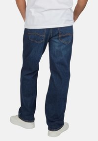 Hombre vistiendo jeans de mezclilla azul oscuro y zapatillas blancas, visto desde atrás, con las manos descansando a los lados y una camisa blanca parcialmente visible.