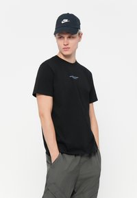 Zwarte katoenen t-shirt met korte mouwen, met blauwe tekst aan de voorkant. Draagt met grijze cargobroek en een donkere baseballcap.