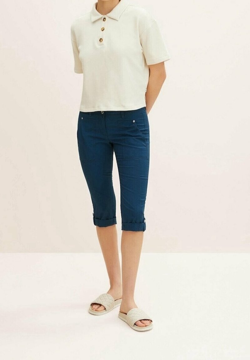 Femme portant un polo crème à manches courtes, un pantalon capri bleu foncé retroussé et des sandales crème à enfiler, debout devant un fond uni.