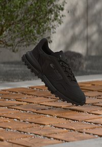 Baskets noires Lacoste avec semelle texturée flottant au-dessus de carreaux en bois et pierre en extérieur, avec un fond flou de verdure et de mur.