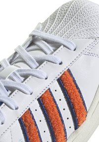 Bílá teniska s texturovaným svrškem, zdobená třemi oranžovými chlupatými pruhy ohraničenými modrým šitím. Šňůrky jsou světle šedé; podrážka je krémová.