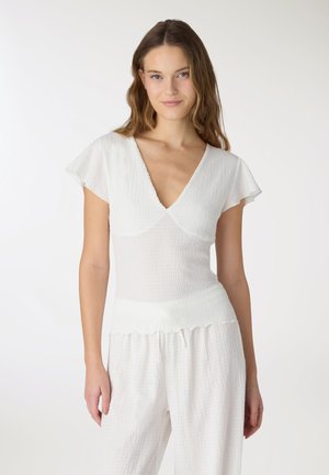 Giovane donna con capelli lunghi che indossa una maglietta bianca a V testurizzata e pantaloni bianchi larghi, in piedi davanti a uno sfondo chiaro e uniforme.