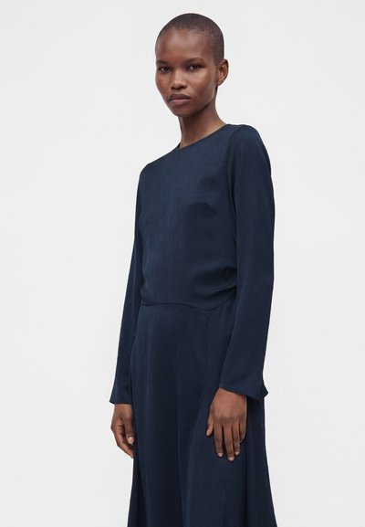 Femme aux cheveux courts portant une robe bleu foncé à manches longues, se tenant devant un fond clair uni.
