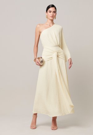 Vrouw draagt een gestructureerde crème kleurige one-shoulder jurk, houdt een kleine clutch vast, en is versierd met gouden oorbellen, armbanden en sandalen met bandjes.