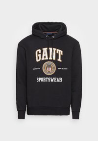 Černá mikina s kapucí na šňůrku, s potiskem "GANT" a "SPORTSWEAR" v bílé a zlaté barvě. Měkký materiál, žebrované manžety a spodní lem.
