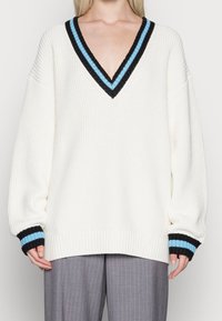 Pull en tricot blanc avec un col en V, présentant un design de rayures noires et bleues au niveau du col et des poignets. Manches longues à coupe ample.