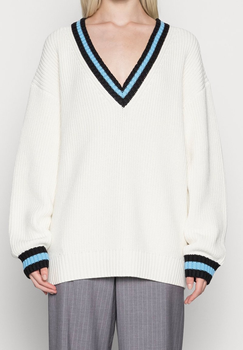 Pull en tricot blanc avec un col en V, présentant un design de rayures noires et bleues au niveau du col et des poignets. Manches longues à coupe ample.