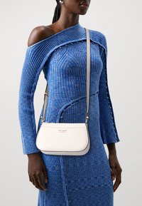 White leather crossbody bag with a curved shape, top zipper, and shoulder strap. Features a gold logo and contrasting trim.