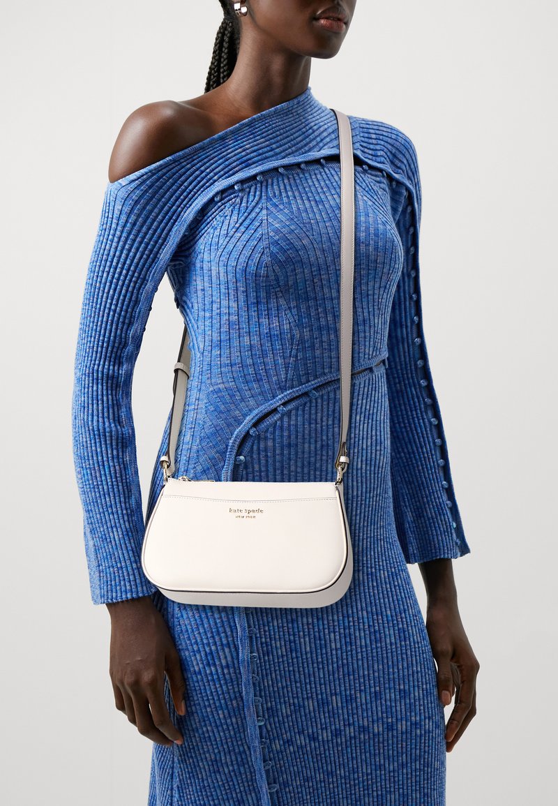 White leather crossbody bag with a curved shape, top zipper, and shoulder strap. Features a gold logo and contrasting trim.