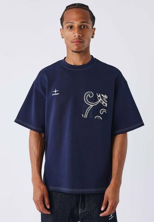Jonge man met gevlochten haar, die een marineblauw oversized t-shirt draagt met een gemarmerde zak en een klein wit logo op de borst.