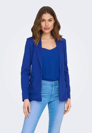 Blå blazer med et struktureret design, der har klapkraver og frontlommer, lagdelt over en matchende blå top og lyseblå jeans.