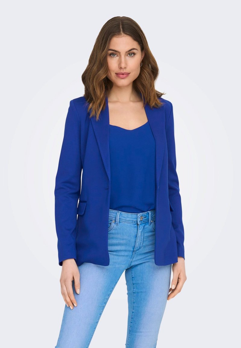 Blazer azul con un diseño estructurado, que presenta solapas de pico y bolsillos delanteros, superpuesto a una blusa azul a juego y jeans azul claro.
