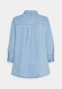 Camisa de denim azul claro de manga larga con puños abotonados, costura del yugo trasero y pliegue central en la parte posterior, exhibida sobre un fondo blanco.