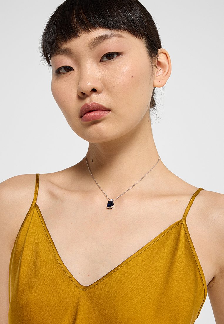 Silberne Halskette mit einem rechteckigen blauen Anhänger, getragen von einem Modell mit einem senfgelben Satinoberteil, das das Design des Accessories hervorhebt.