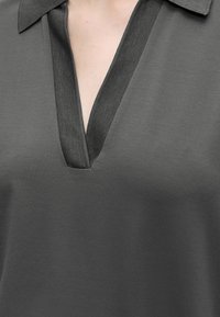 Gros plan d'une chemise grise à col avec un profond col en V portée par une personne, montrant le haut de la poitrine et les détails du col.