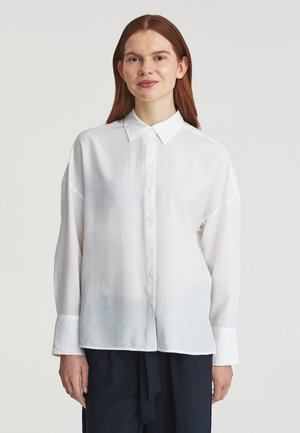 Femme aux longs cheveux roux portant une chemise blanche ample à boutons et un pantalon foncé, debout devant un fond clair uni.